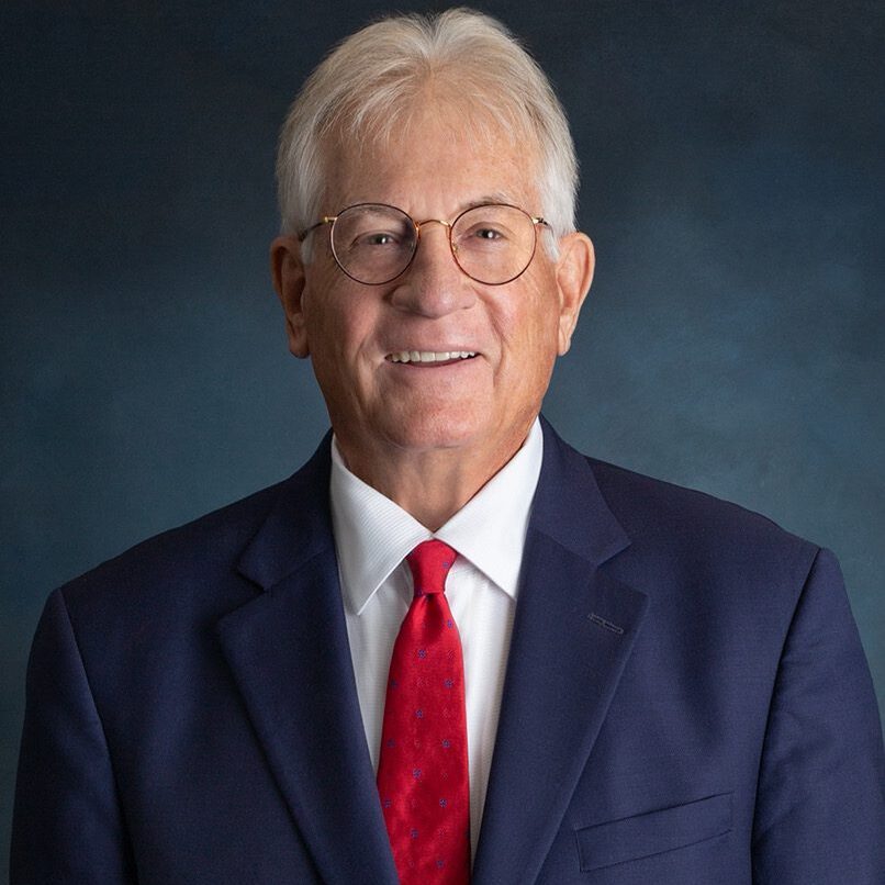 Elderly man in a suit and red tie smiling against a dark background.